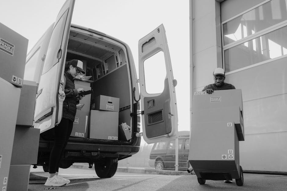 A man wearing a dark jacket, black pants, and a baseball cap is loading or unloading a white panel van with open rear doors, filled with several cardboard boxes of various sizes, some sealed with pack